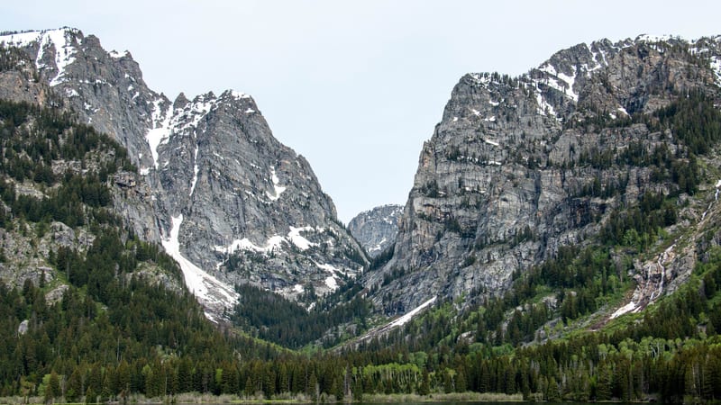 Two backpackers ascend a rocky alpine trail in Grand Teton National Park, with jagged snow-patched peaks rising sharply above a deep forested canyon under a …
