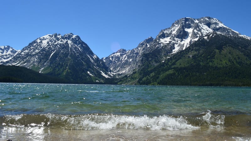 A glacially fed alpine lake with small waves lapping a sandy shore in the foreground, backed by snow-capped Teton peaks and dense conifer forests under a cle…
