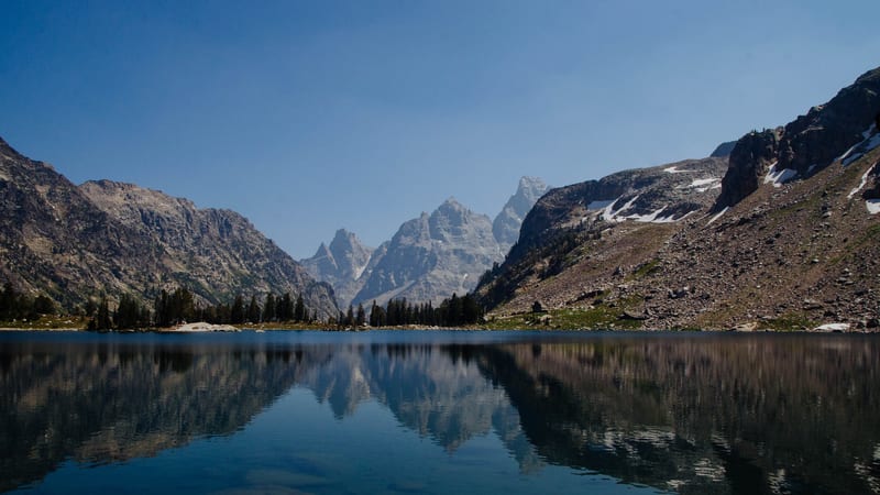 A calm alpine lake reflects the jagged, snow-streaked peaks of the Teton Range under a clear blue sky, framed by rocky canyon walls and a band of conifer tre…
