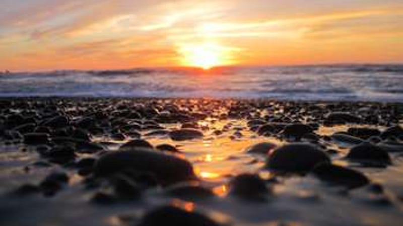 A low-angle ground-level view of a rocky pebble-strewn beach at sunset, with warm orange and golden light reflecting off wet stones and breaking surf.