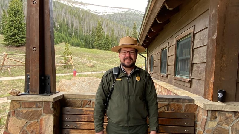 A National Park Service ranger stands behind an information table loaded with Junior Ranger booklets and educational materials, set on a covered stone porch …
