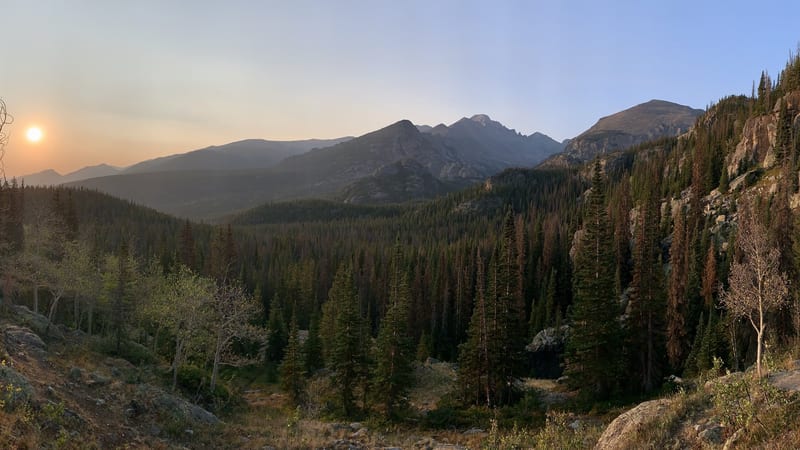 A panoramic view of a forested mountain valley in Rocky Mountain National Park at sunset, with hazy smoke-filtered golden light illuminating conifer-covered …