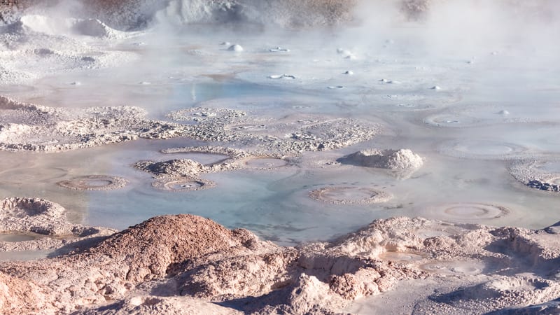 A close-up view of a hydrothermal mud pot or hot spring field featuring boiling bubbles, mineral-encrusted mounds, and milky blue-gray thermal water with ste…