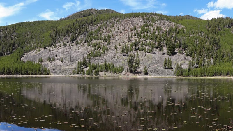 A calm mountain lake with lily pads floating on the surface reflects a rocky, conifer-covered hillside under a partly cloudy blue sky.