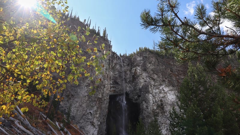 A tall, narrow waterfall cascades down a sheer gray rock cliff face framed by golden-leafed aspen trees on the left and pine branches on the right, suggestin…