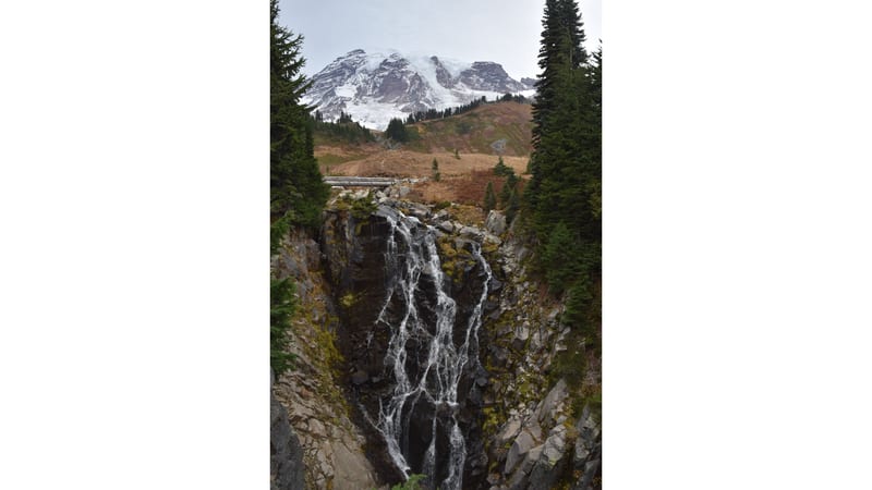 A multi-strand waterfall cascades down a dark basalt rock face at Mount Rainier National Park, with the snow-capped summit and glaciers of Mount Rainier visi…
