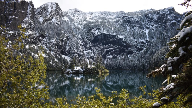 A calm alpine lake reflects snow-dusted rocky cliffs and evergreen forest under overcast winter light.