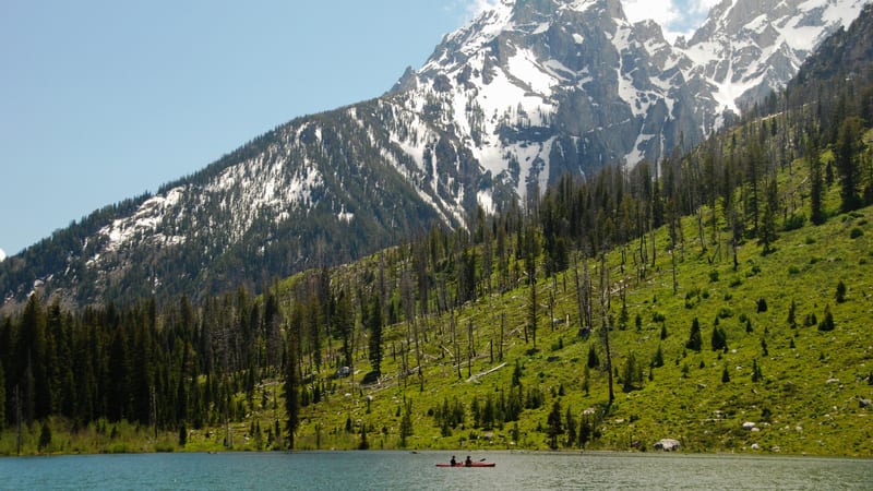 A tranquil alpine lake with vivid turquoise water sits at the base of the jagged, snow-capped Grand Teton peaks in Grand Teton National Park, Wyoming.
