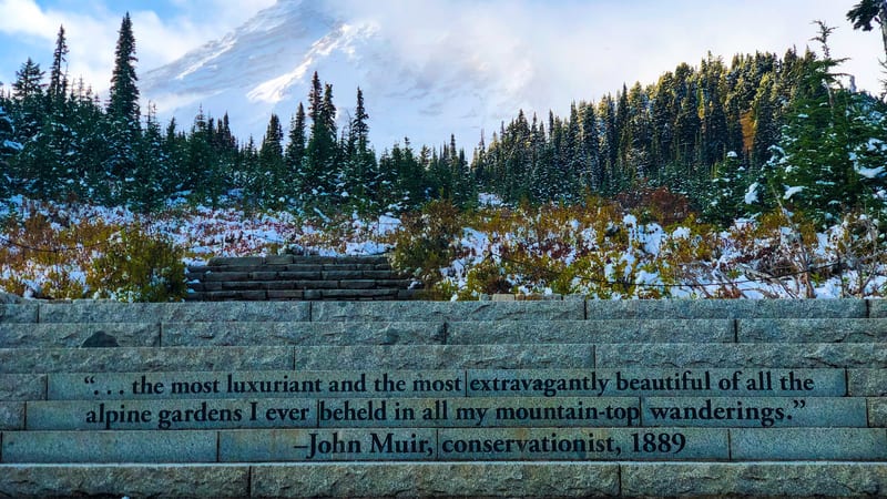A granite retaining wall engraved with an 1889 John Muir quote frames a snow-dusted alpine meadow and conifer forest below the snow-capped peak of Mount Rain…