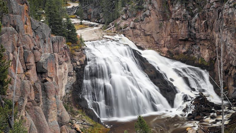 A wide cascade waterfall tumbles over dark volcanic rock through a narrow canyon flanked by rugged reddish-brown cliff walls and dense conifer forest.