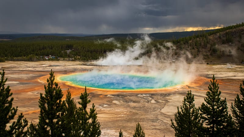 A geyser erupts forcefully at Yellowstone National Park, sending a column of water and steam into the air while a group of tourists watches from a nearby vie…