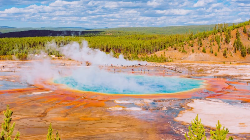 Tourists walk along a raised boardwalk beside the vivid orange and white mineral-encrusted edges of Grand Prismatic Spring at Midway Geyser Basin, with steam…