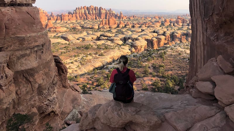A hiker sits on a rocky ledge gazing out over the vast, otherworldly landscape of The Needles district in Canyonlands National Park, with hundreds of red san…