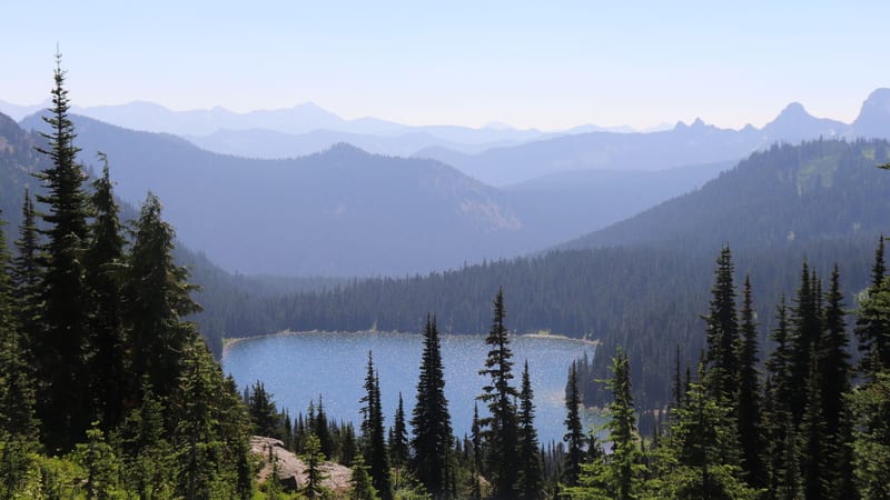 A vivid turquoise alpine lake sits nestled in a forested valley surrounded by dense subalpine fir trees, with layers of hazy blue-green mountain ridges reced…