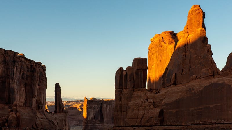 Towering sandstone fins and monoliths line a narrow corridor at Arches National Park, bathed in warm golden-orange sunrise light that illuminates the upper r…