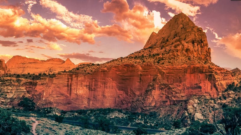 A historic pioneer farmstead with a rustic wooden barn and fenced pastures sits beneath towering red sandstone cliffs in Capitol Reef National Park, Utah.