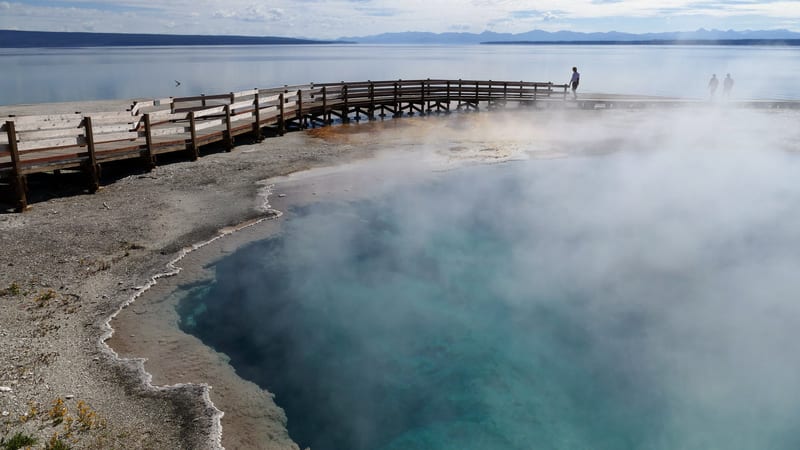 A steaming turquoise hot spring pool along the shore of Yellowstone Lake, viewed from a wooden boardwalk that curves around its edge.