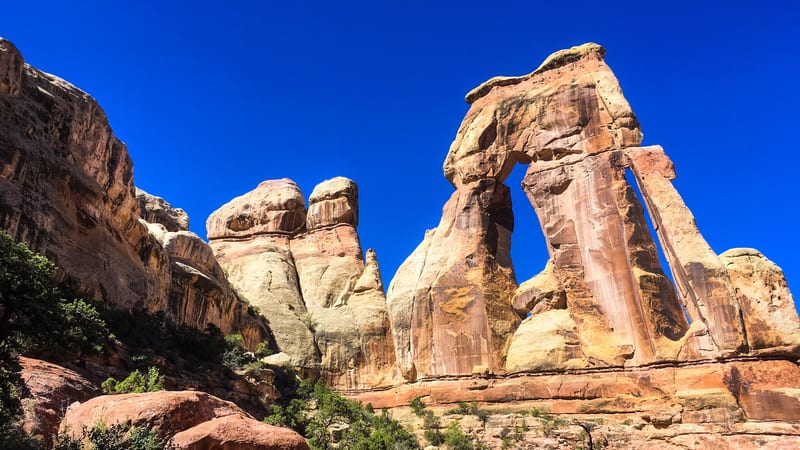 A dramatic sandstone arch rises against a vivid deep-blue sky in what appears to be Canyonlands National Park, with warm orange-red rock formations and scatt…