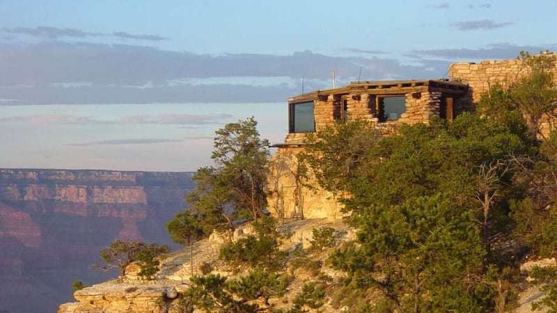 A rustic stone-and-log observation building perches at the edge of the Grand Canyon's South Rim, surrounded by pinyon pine and juniper trees.