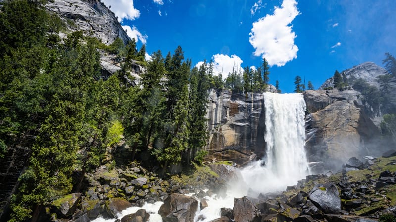 A large waterfall drops vertically down a granite cliff into a rocky river, flanked by evergreen trees and gray granite walls under a blue sky with white clo…