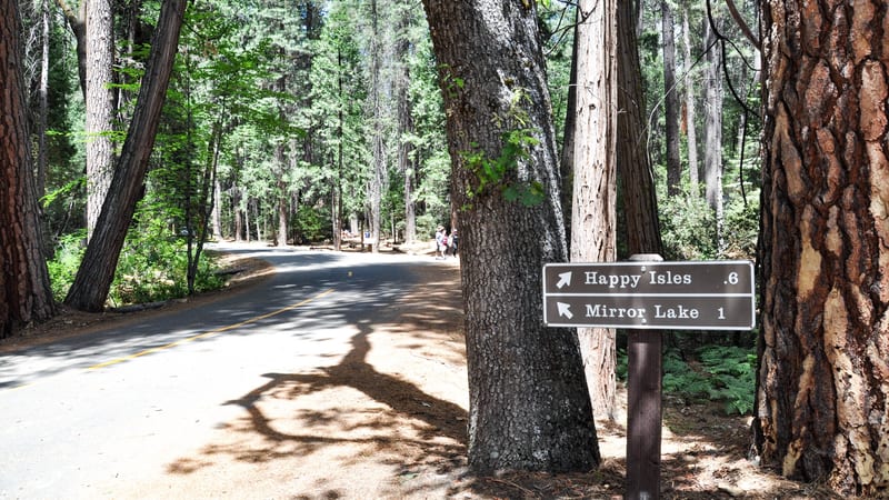 A shaded forest road winds through tall conifers in Yosemite Valley, with a brown National Park Service directional sign pointing toward Happy Isles (0.6 mil…